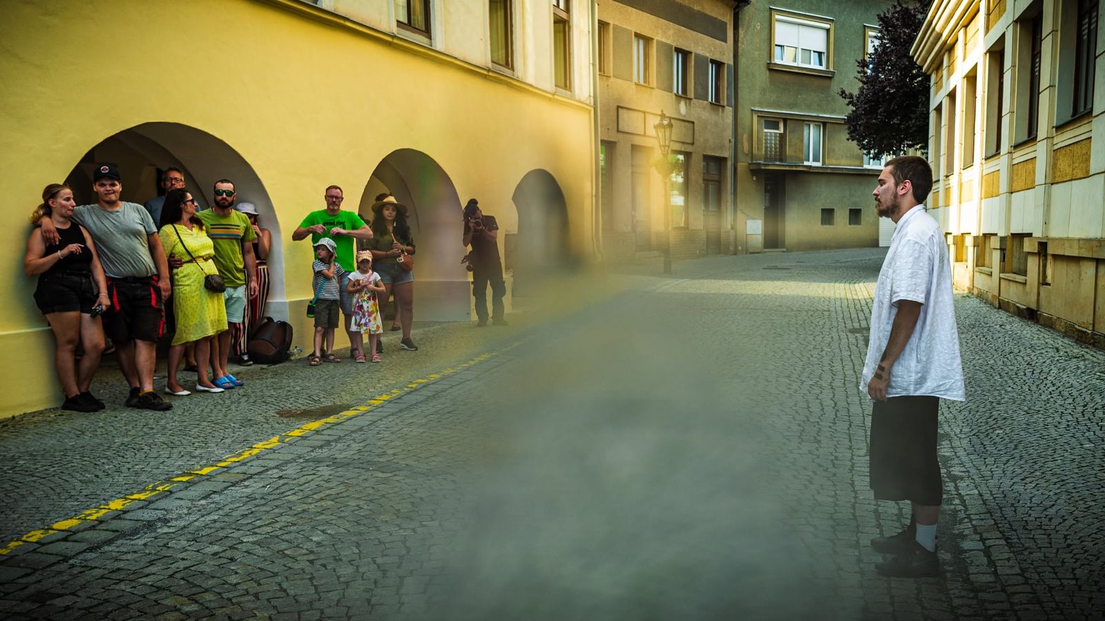 A performer standing in the street in a low mist, guests watching from under the arcade