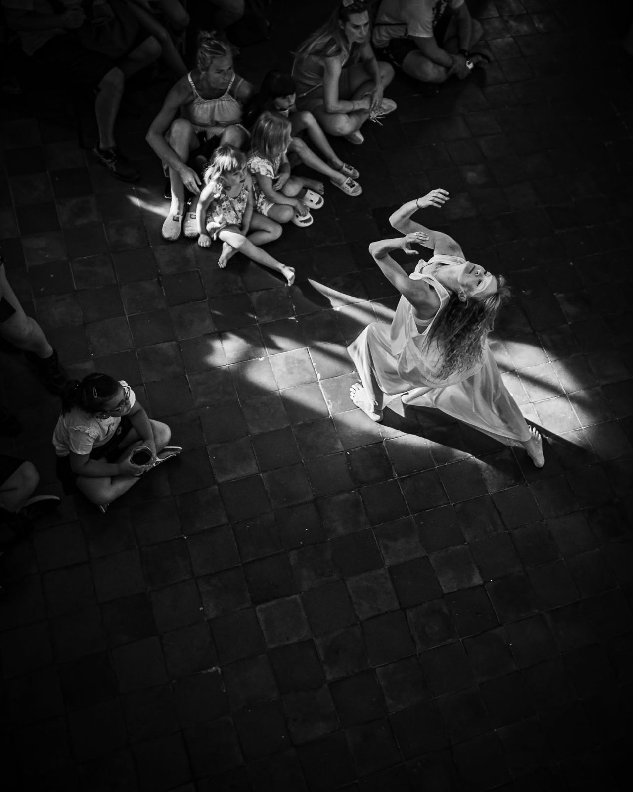 Black and white, from above: a dancer in a light dress turning on the paving, guests seated around her