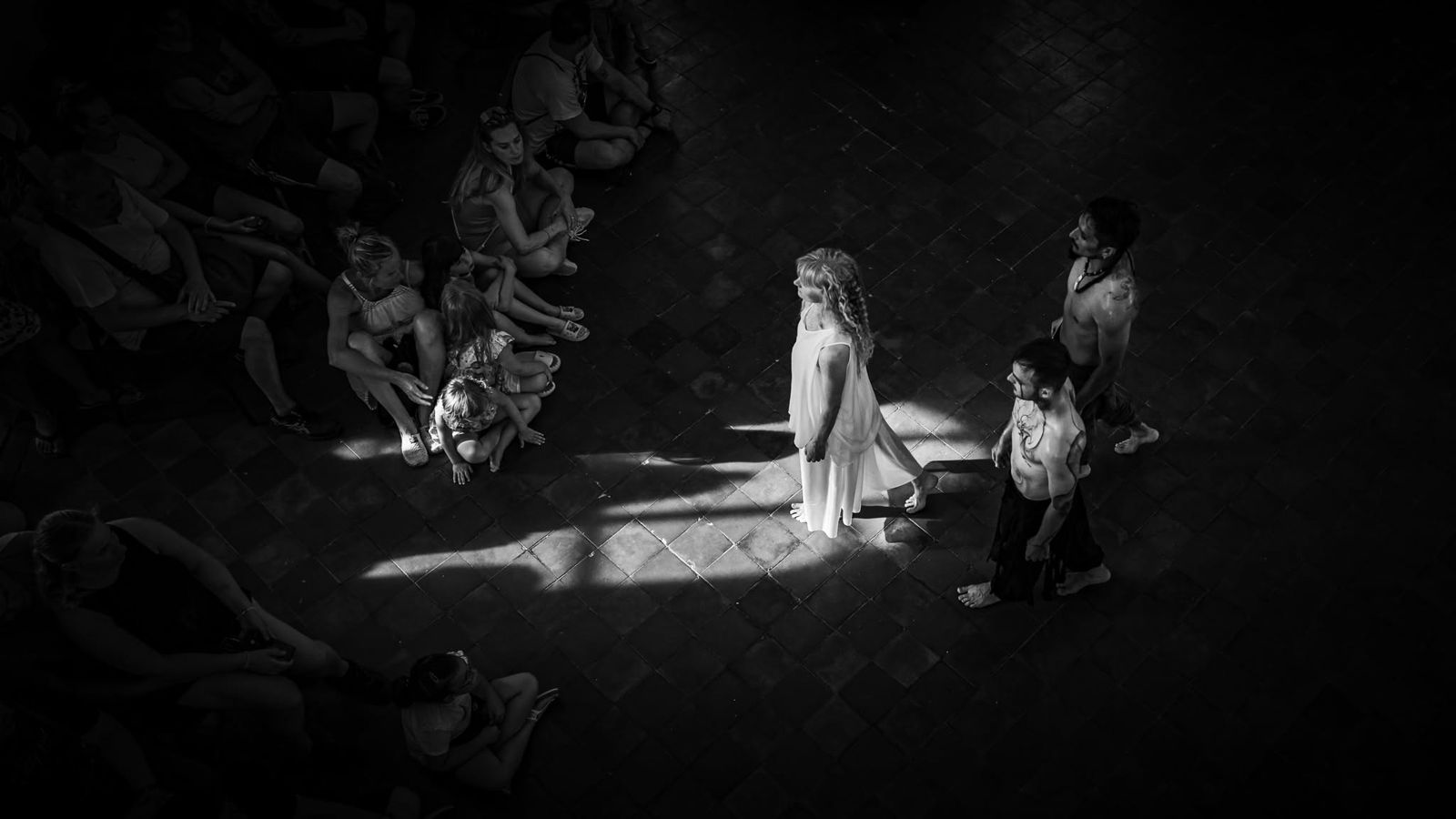 Black and white: a girl in a white dress in a patch of light on the stones, children around her