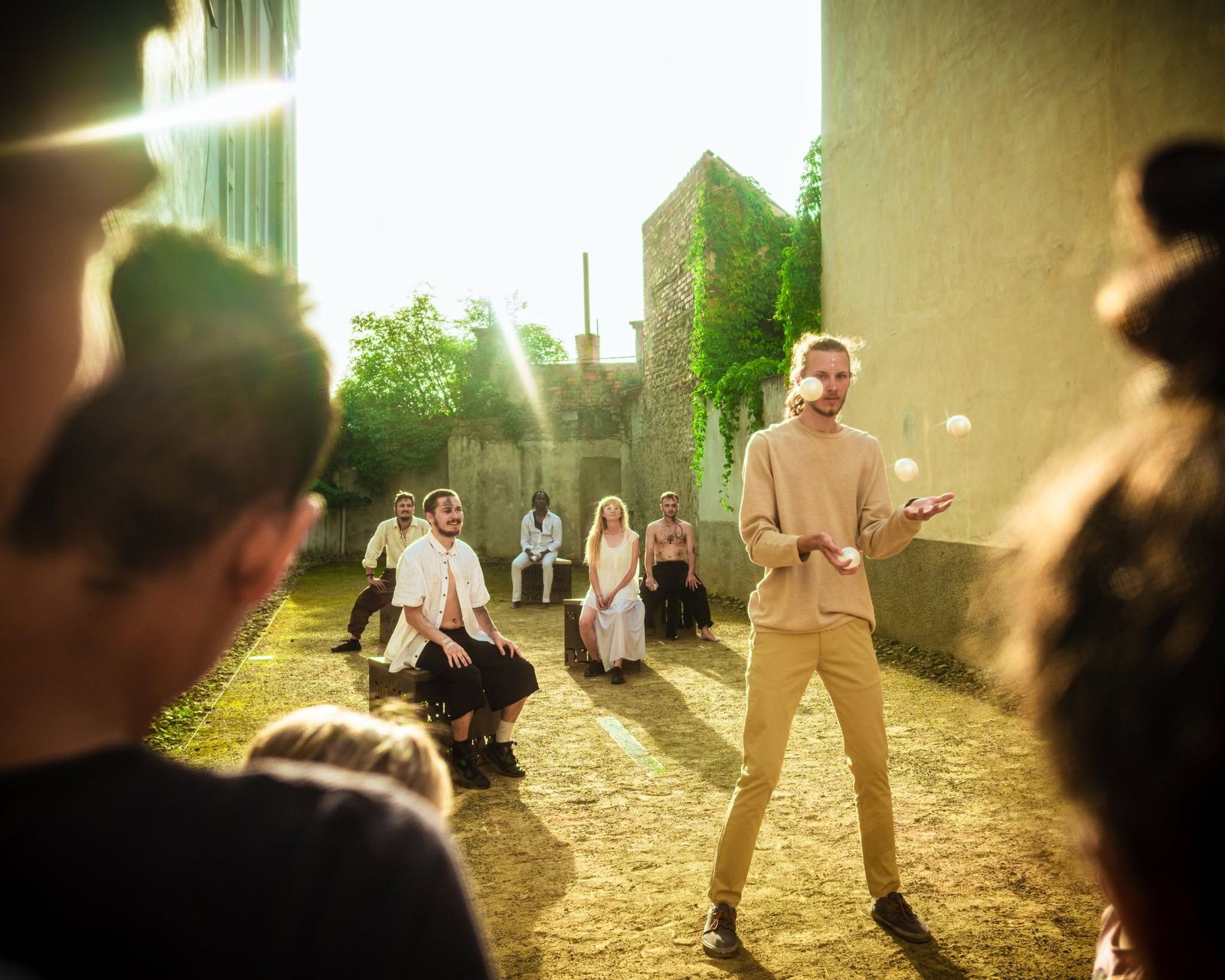 A man in a beige suit juggling in a sunlit garden, other performers seated in a circle behind