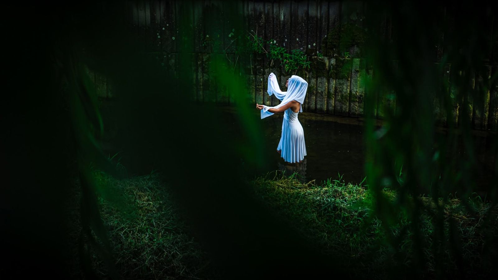 A figure in a white hood and dress standing in the dark green of a garden