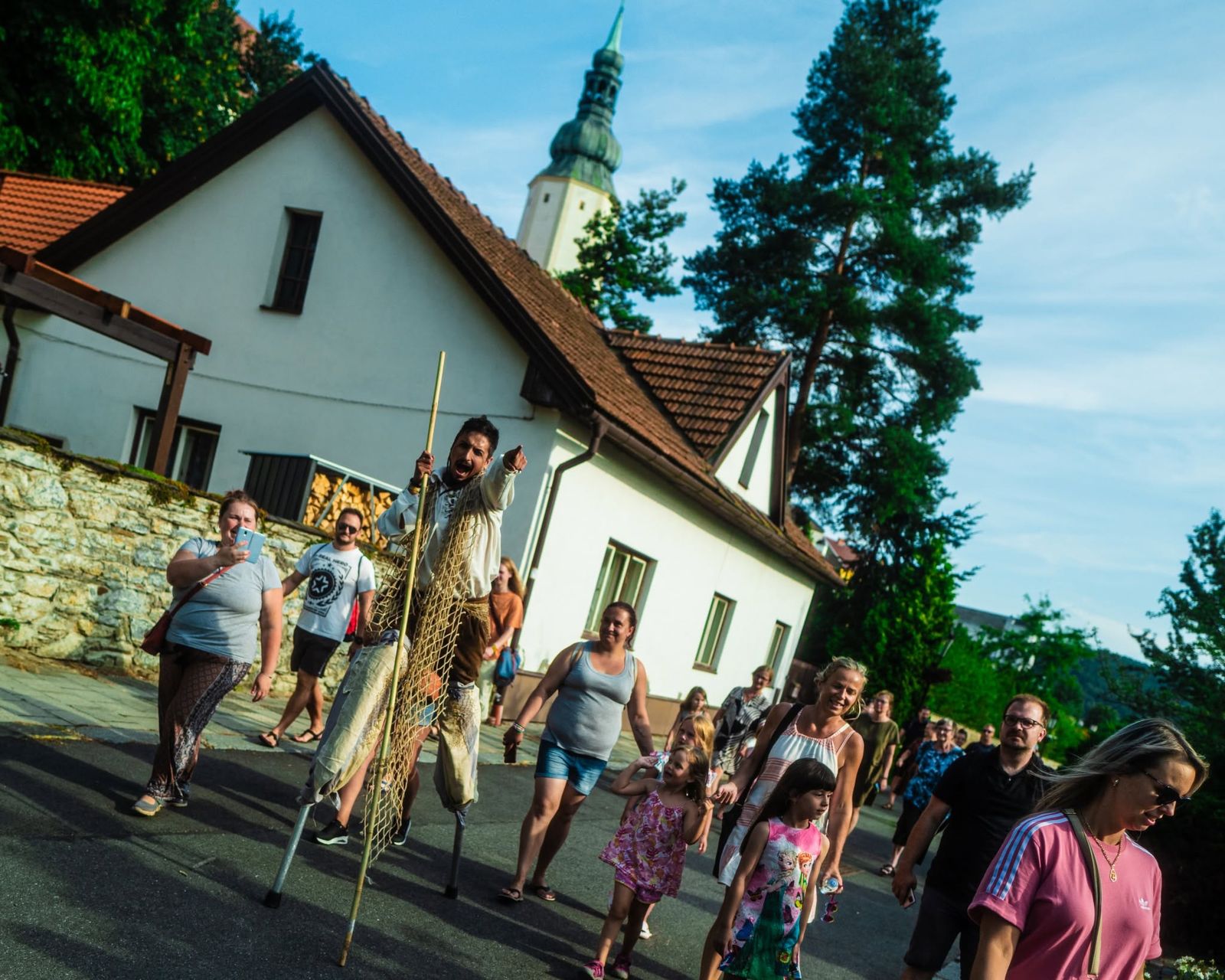 A performer on stilts leading families down a village street, a church tower behind the trees