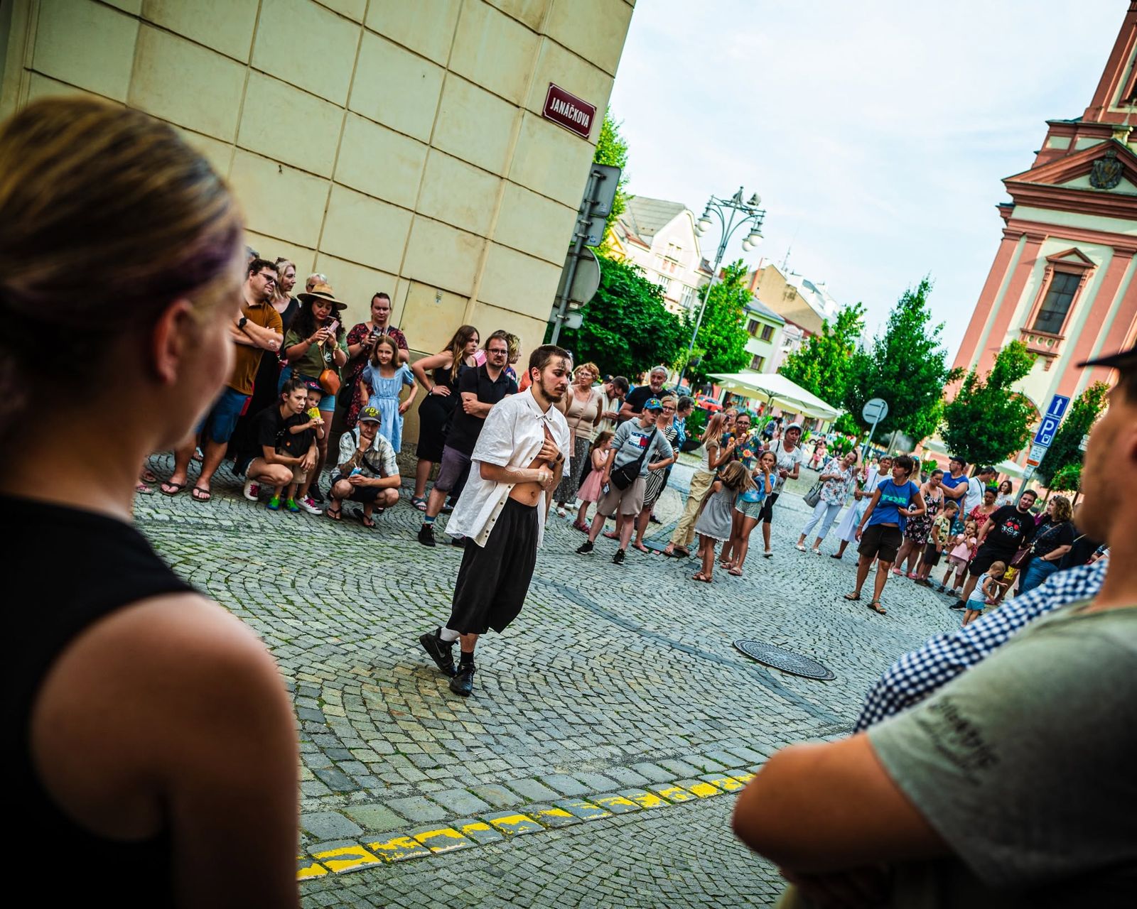 A dancer in a white shirt moving across the cobbled street, a crowd of guests in a wide circle around him