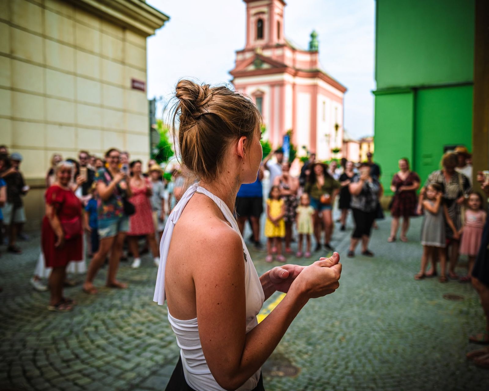 A dancer with her back to the camera opens her hands to the crowd on the street, a pink church tower behind