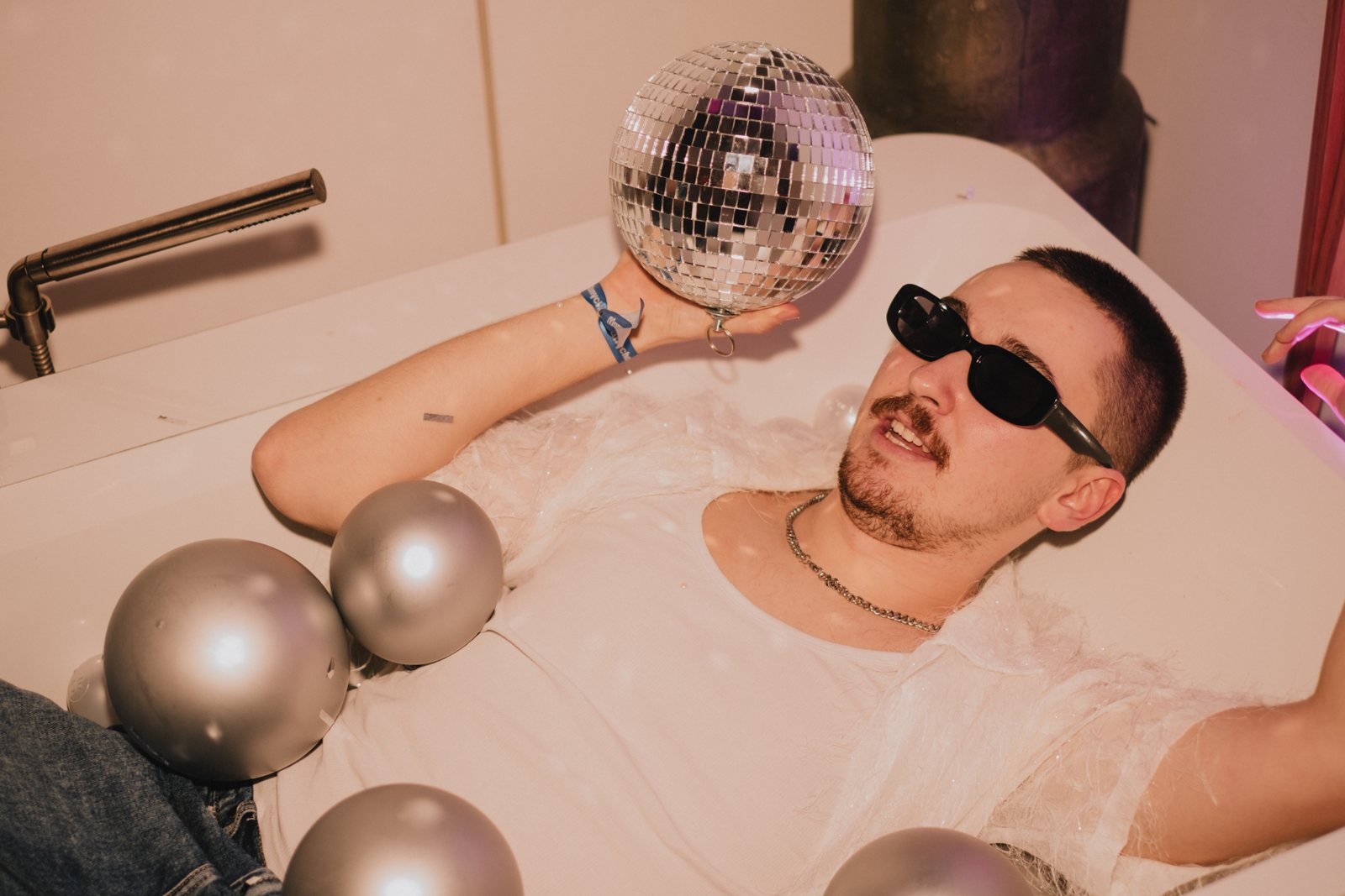 A guest lying in a bathtub full of silver balls, holding a disco ball
