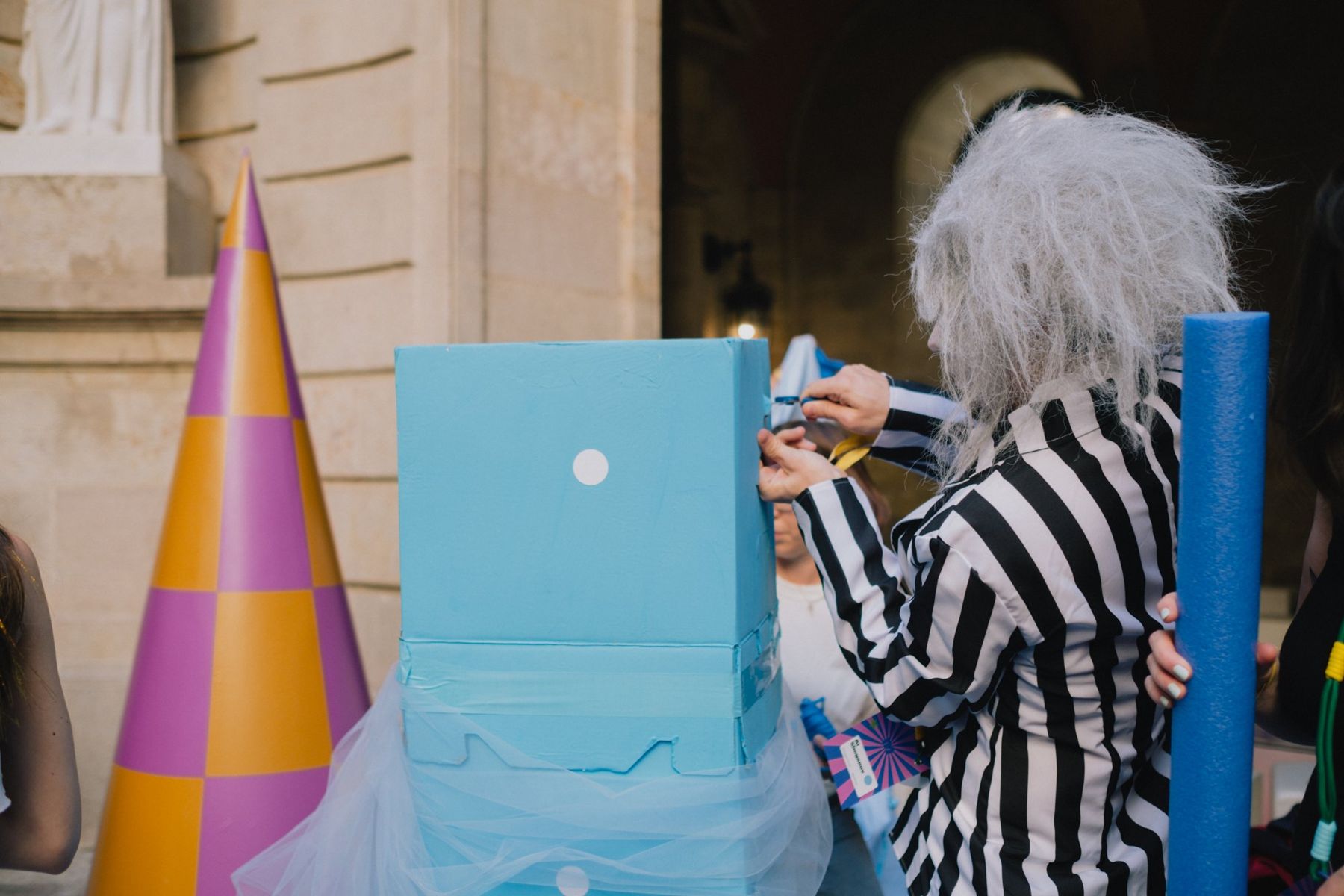 A guest in a striped suit finishing a sky-blue box creature next to a purple and orange cone