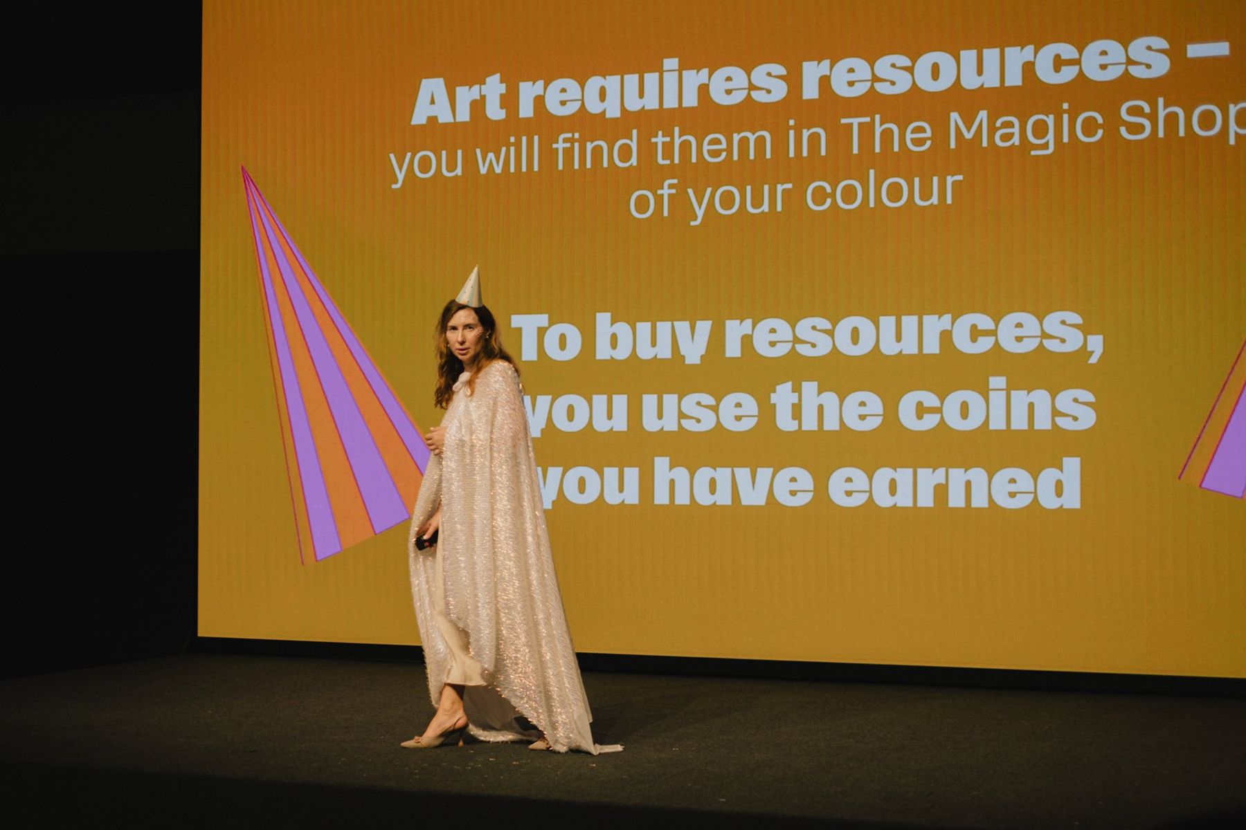 The host on stage in front of a yellow slide explaining that coins buy resources