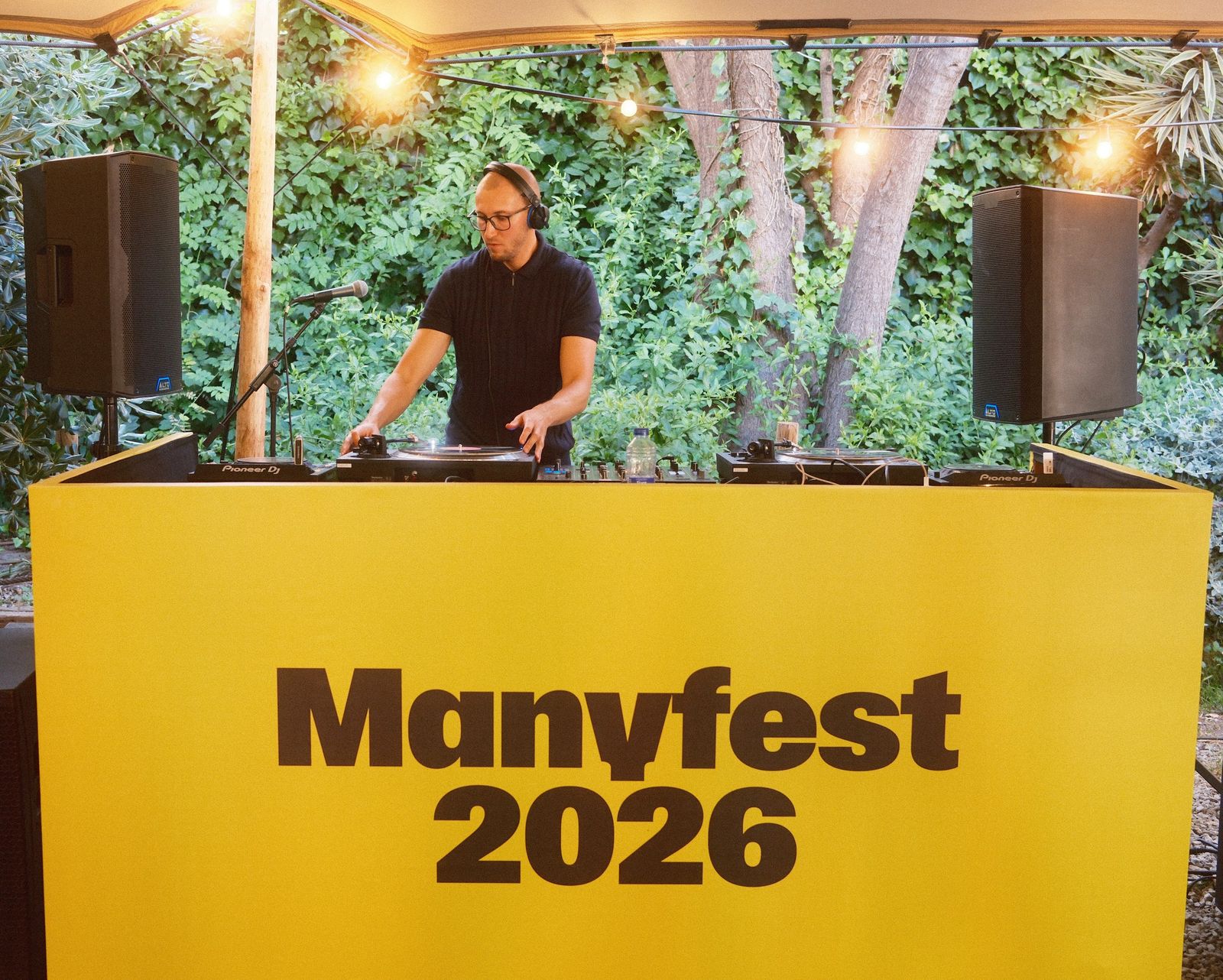 A DJ behind a yellow Manyfest 2026 booth among palm trees
