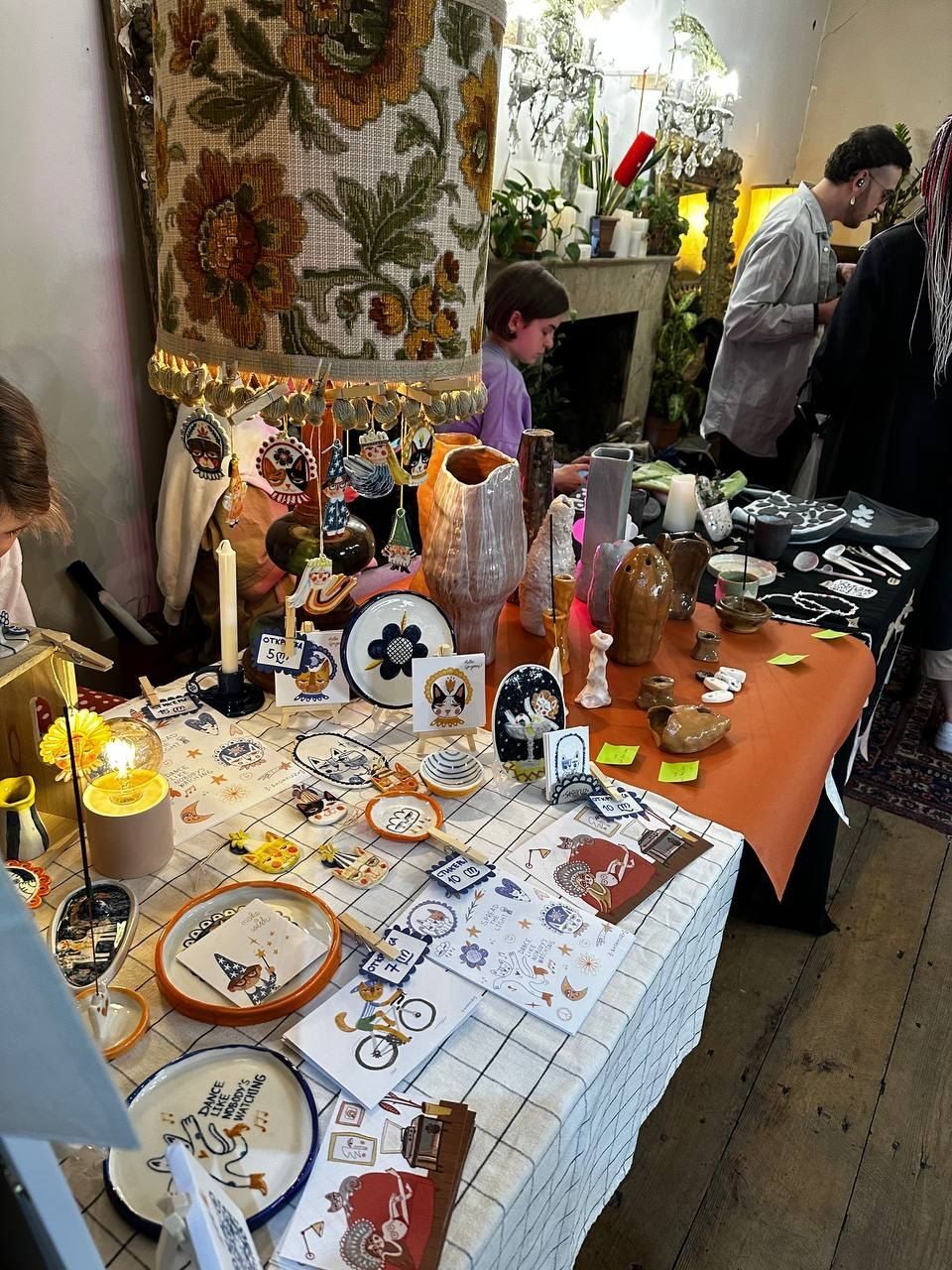 A table of ceramics, plates and prints under an embroidered lampshade