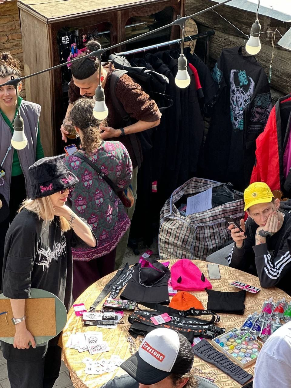 Guests around a round table of caps, beanies and lanyards, black clothes on the rail behind